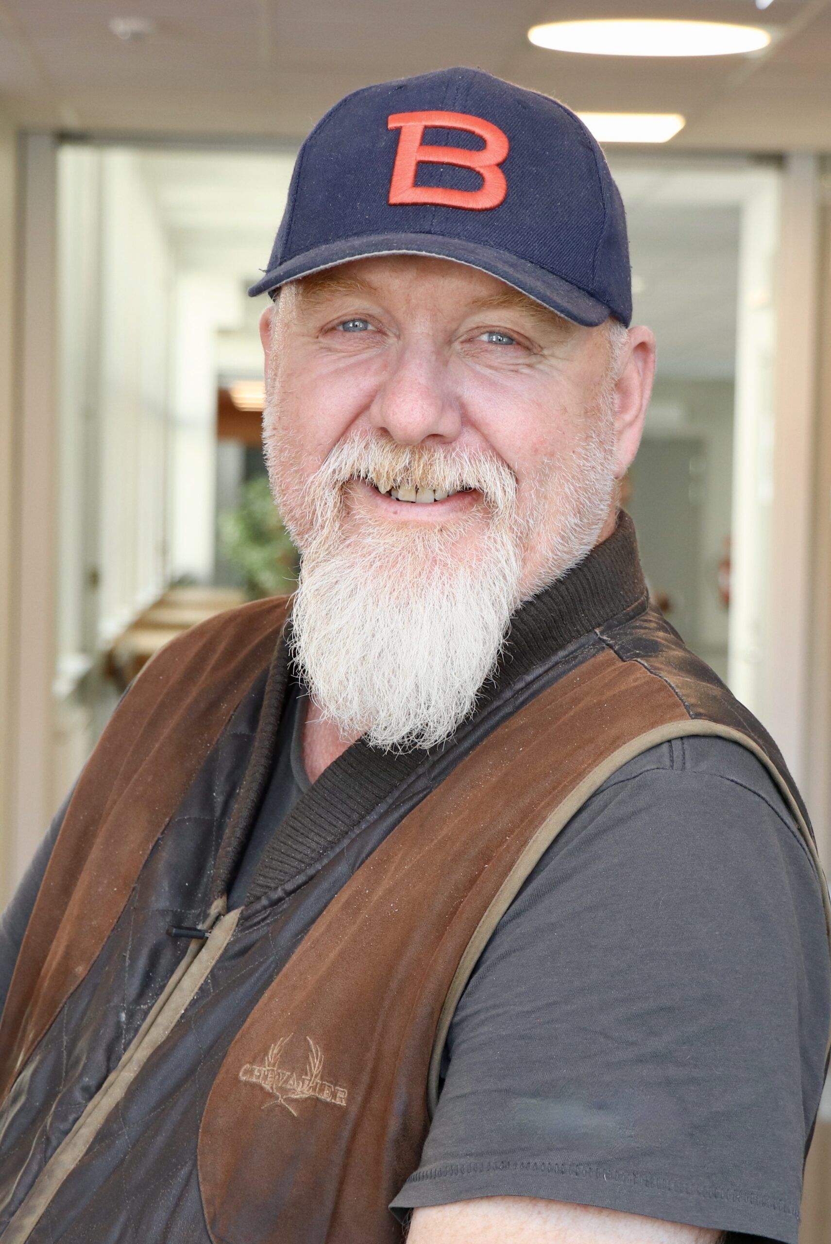 En man i en grå tröja och brun väst och en blå Bergstrands keps som ler.