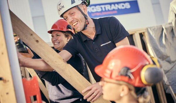 En grupp byggnadsarbetare, klädda i hjälm och säkerhetsutrustning, skrattar och samarbetar på en träkonstruktion.