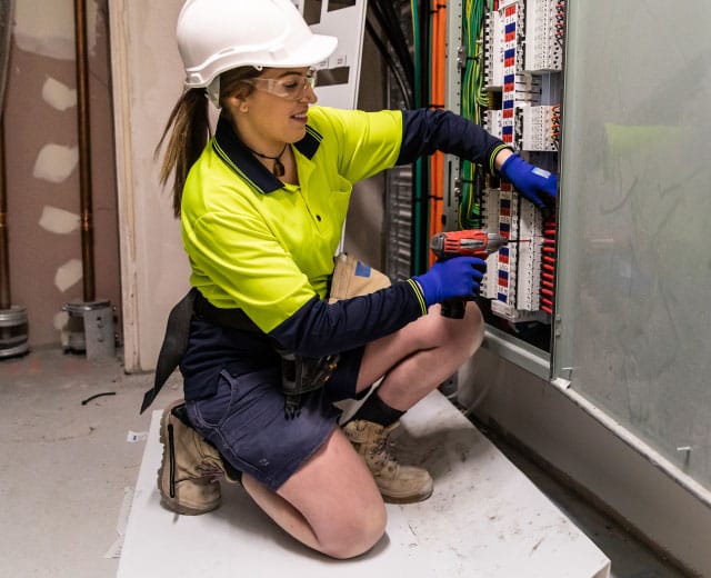 Elektriker i synlig skjorta och hjälm knäböjer medan han använder en borr för att arbeta på en elektrisk panel.