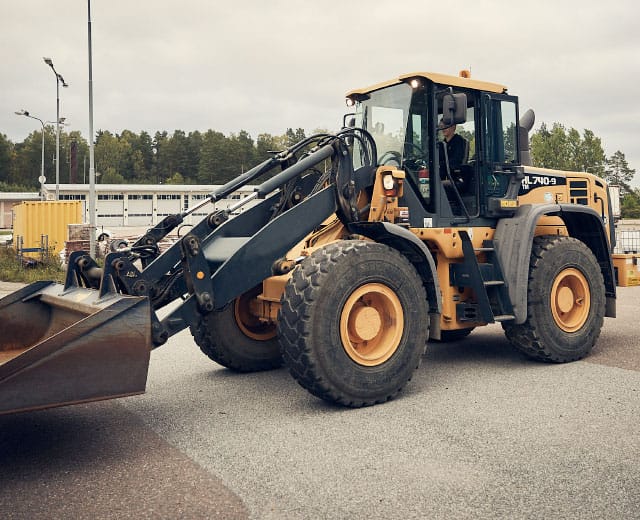 En gul frontlastare står parkerad på en industritomt med träd och byggnader i bakgrunden.