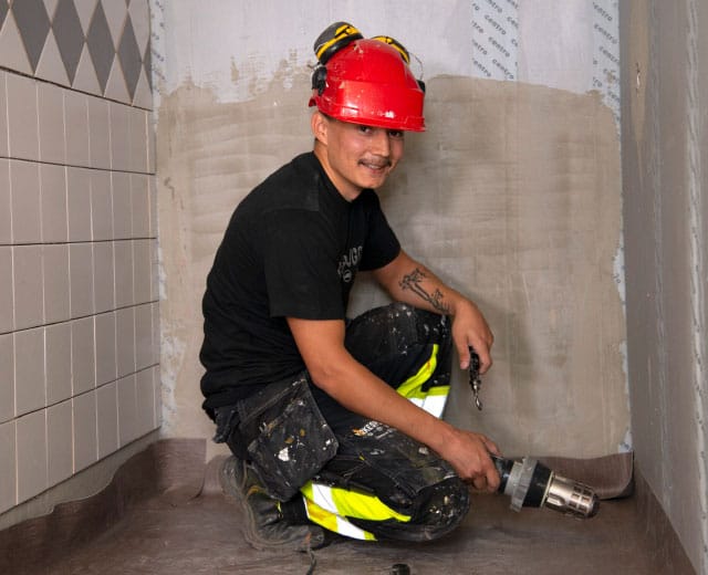 En person som bär en röd hjälm och svart skjorta knäböjer på golvet och håller i en borr i ett kaklat rum under renovering.
