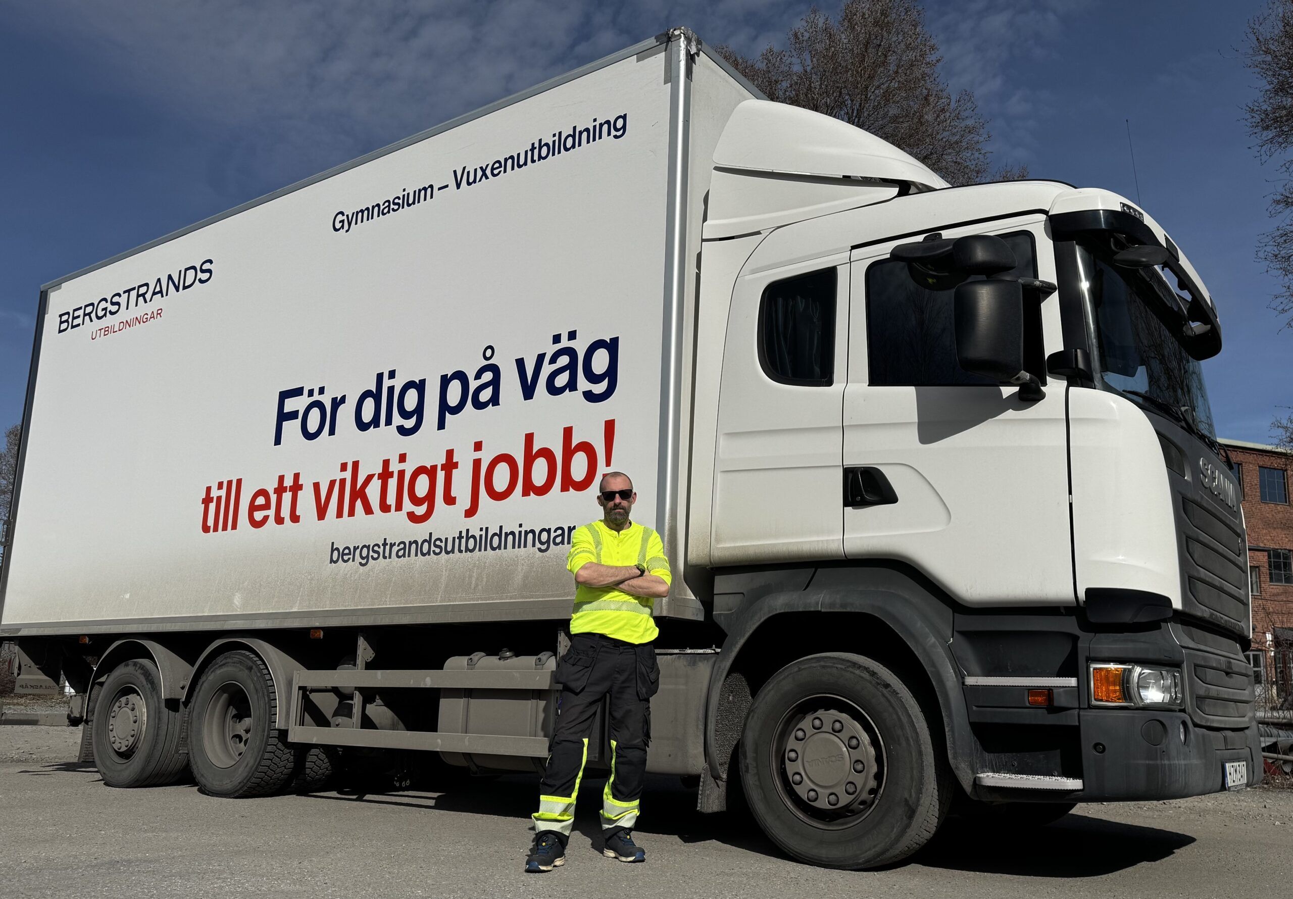 En man i en gul synlig jacka står framför en stor vit lastbil med svensk text som främjar utbildning och arbetsträning.