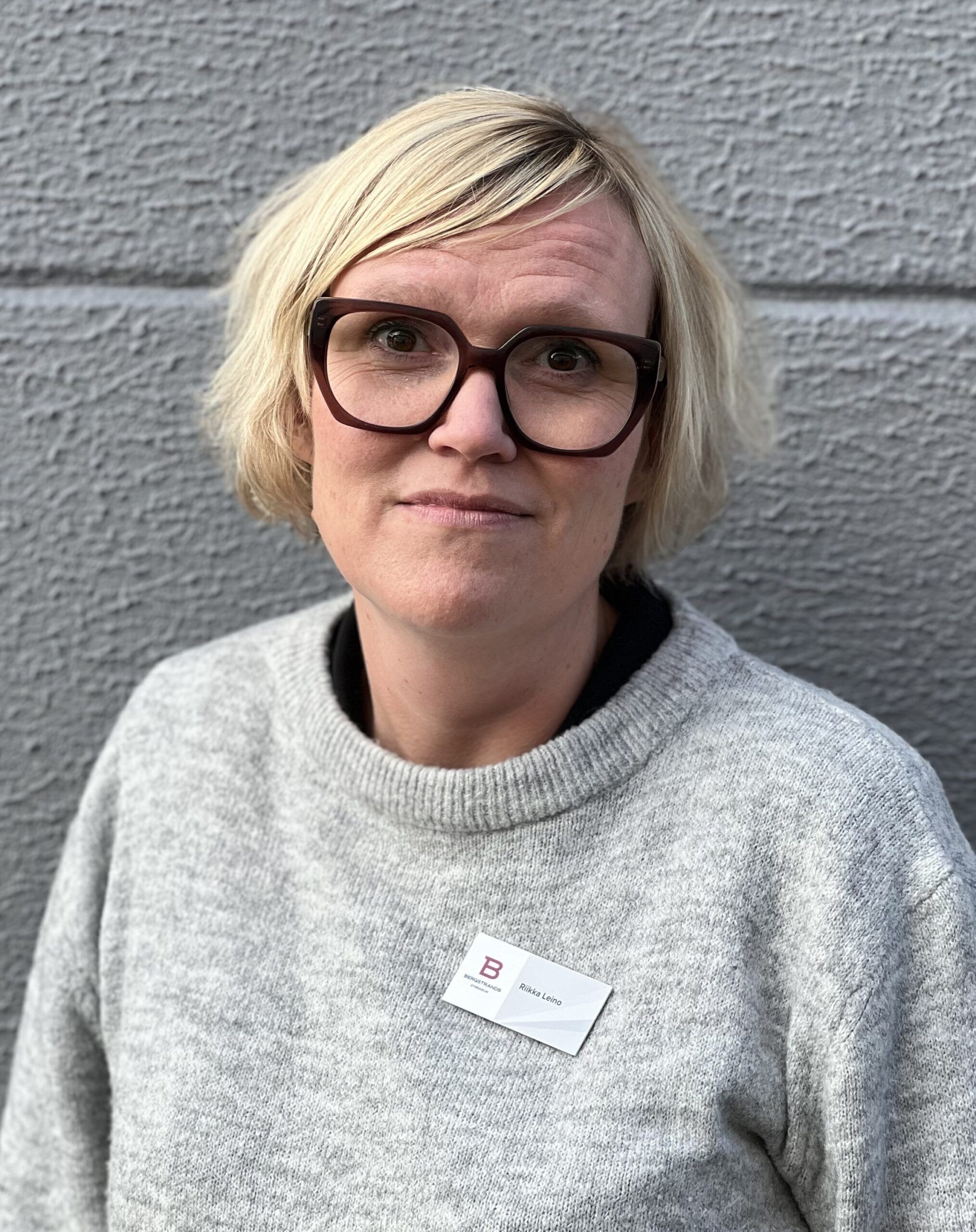 En person med kort blont hår och stora glasögon, klädd i en grå tröja och en namnskylt står framför en grå vägg.