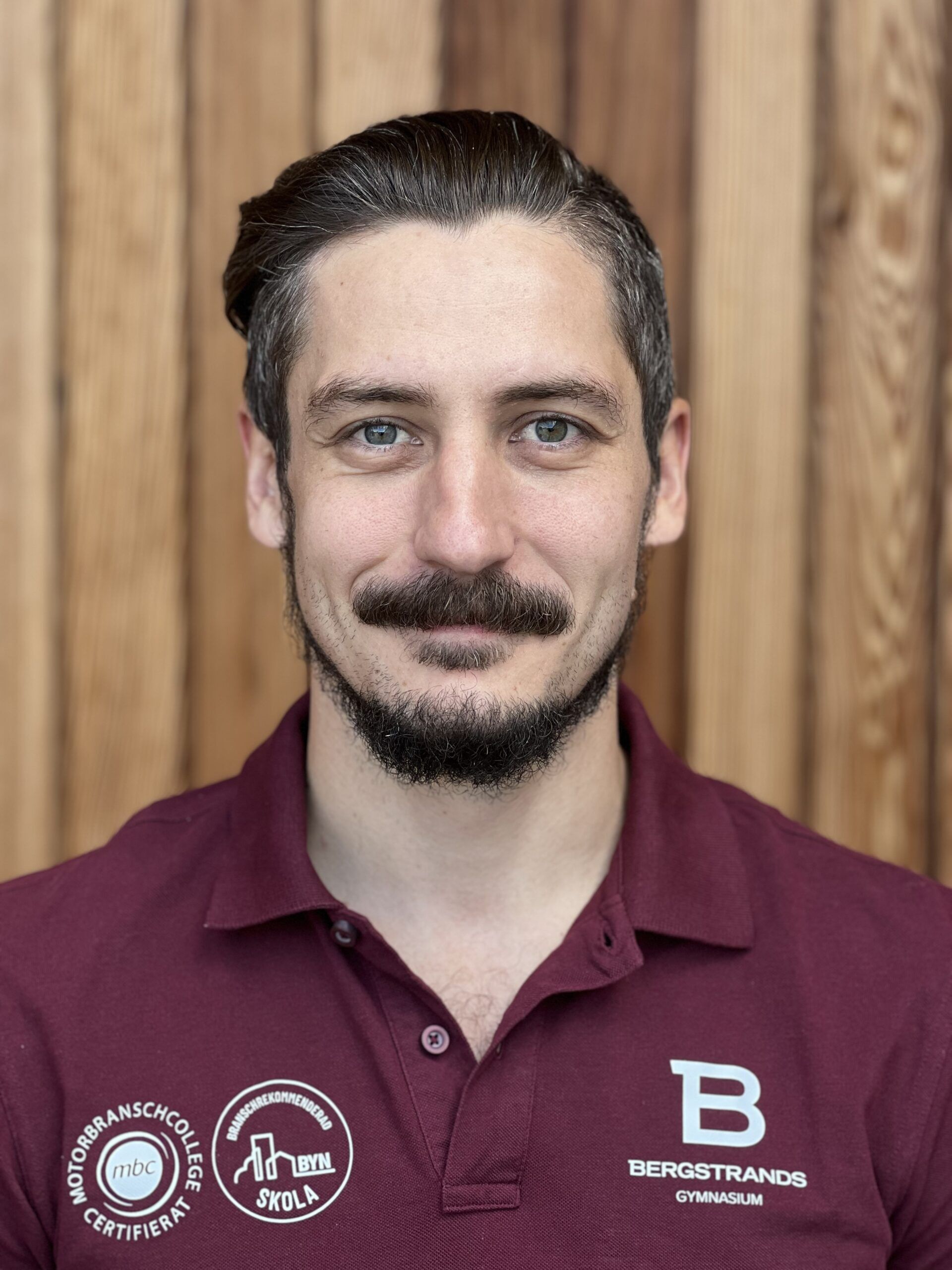 En man med brunt hår och mustasch, iklädd en rödbrun polotröja med Bergstrands Gymnasium och andra logotyper, står framför en trävägg.