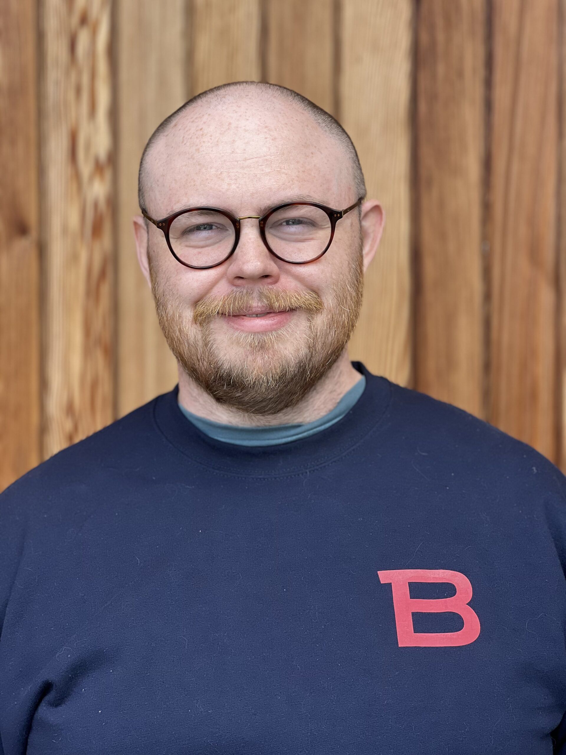 En person med glasögon och skägg står framför en trävägg, iklädd en marinblå sweatshirt med ett stort rött "B" på.