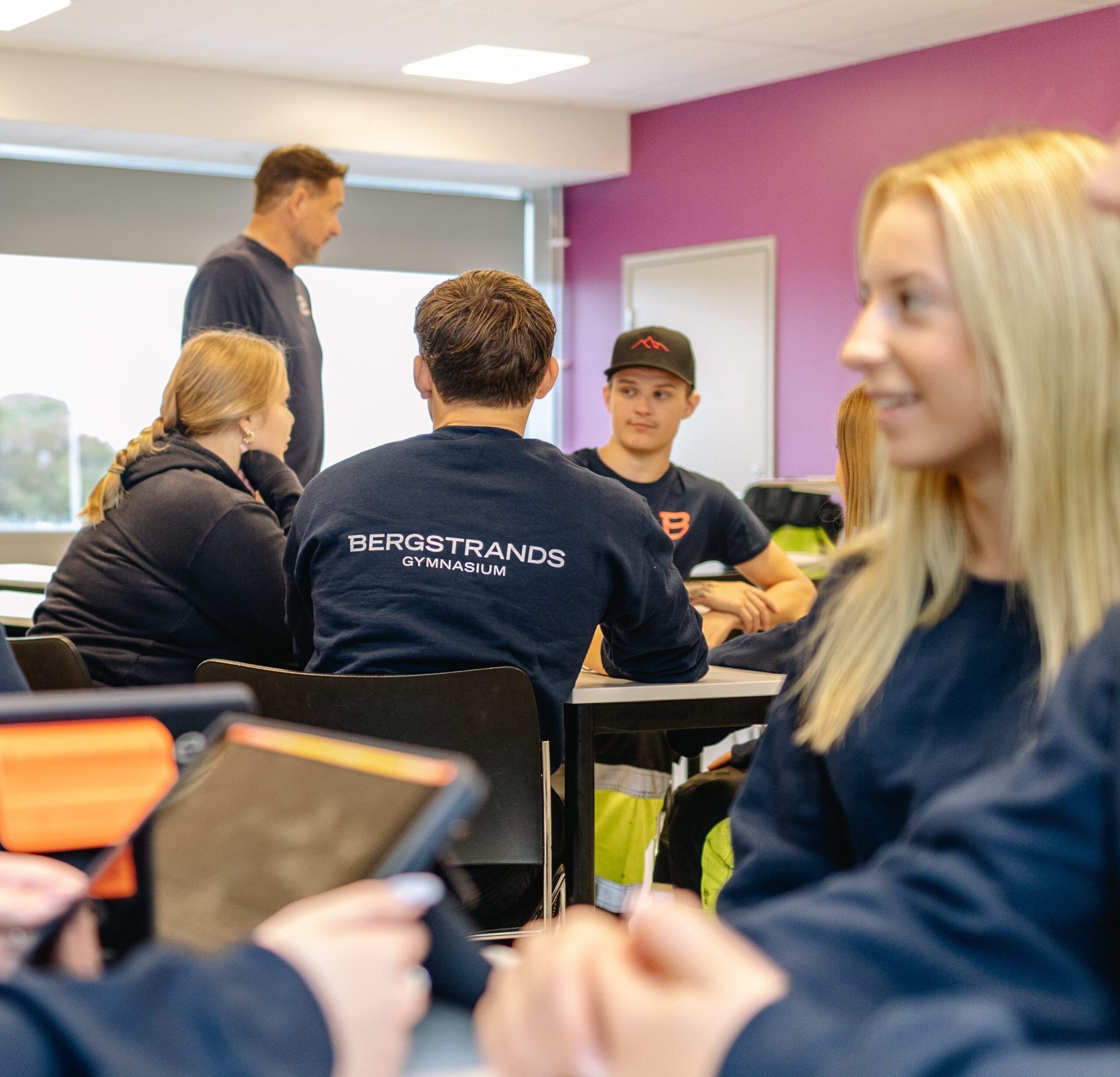 Elever sitter vid bord i ett klassrum, varav en bär en tröja med texten "Bergstrands Gymnasium". En lärare står vid ett fönster och några elever använder surfplattor eller anteckningsböcker.